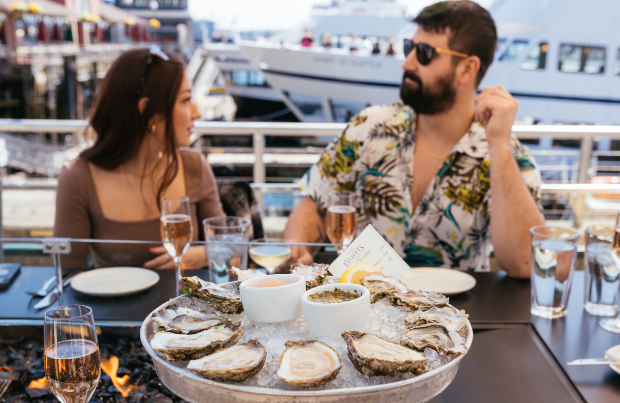 A man and woman sit at an outdoor restaurant table by the water, with a large tray of oysters and drinks in front of them. A docked boat and other diners are visible in the background.