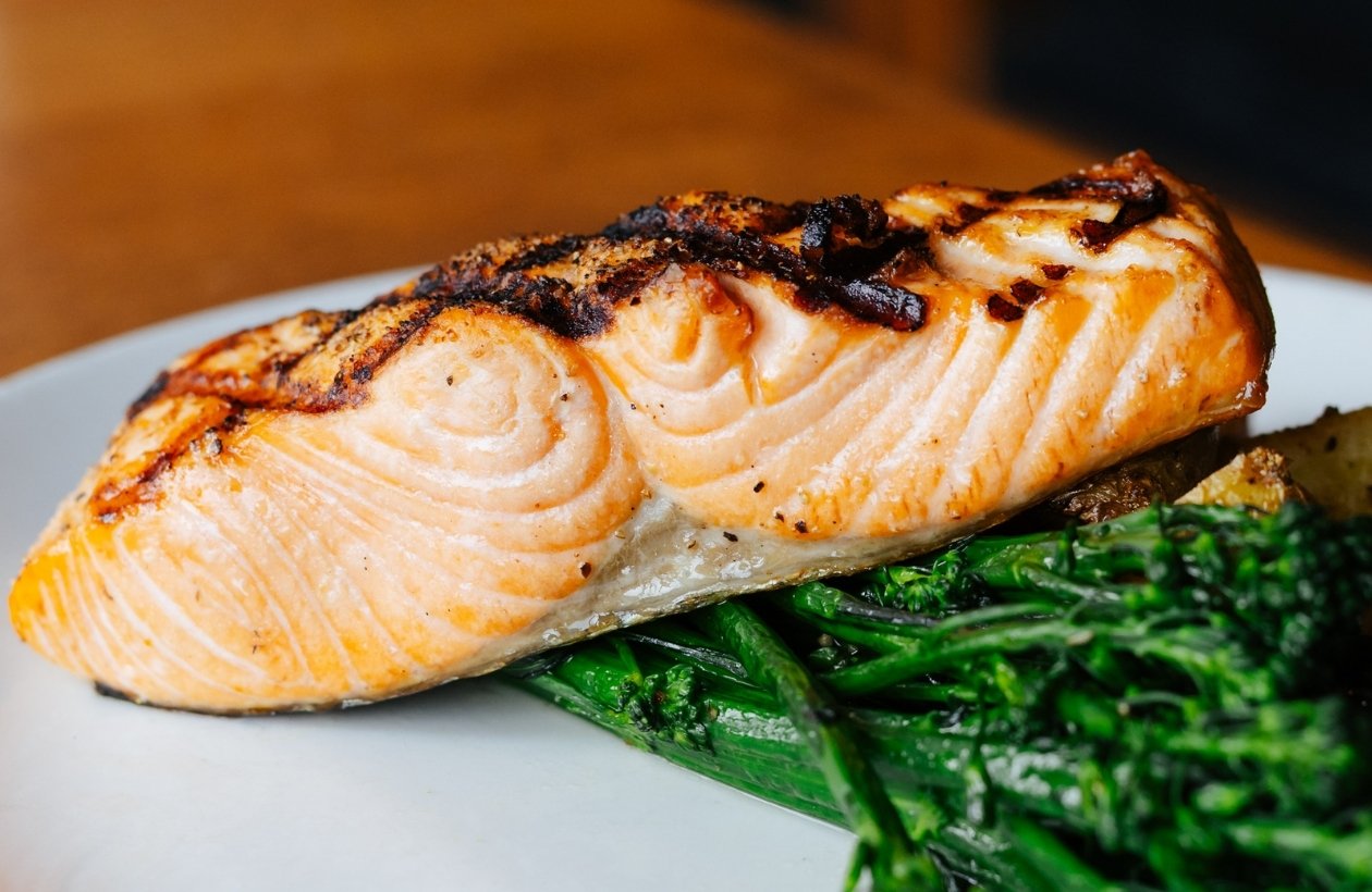 A grilled salmon fillet rests on a bed of sautéed green vegetables, likely broccolini, on a white plate with a blurred brown background.