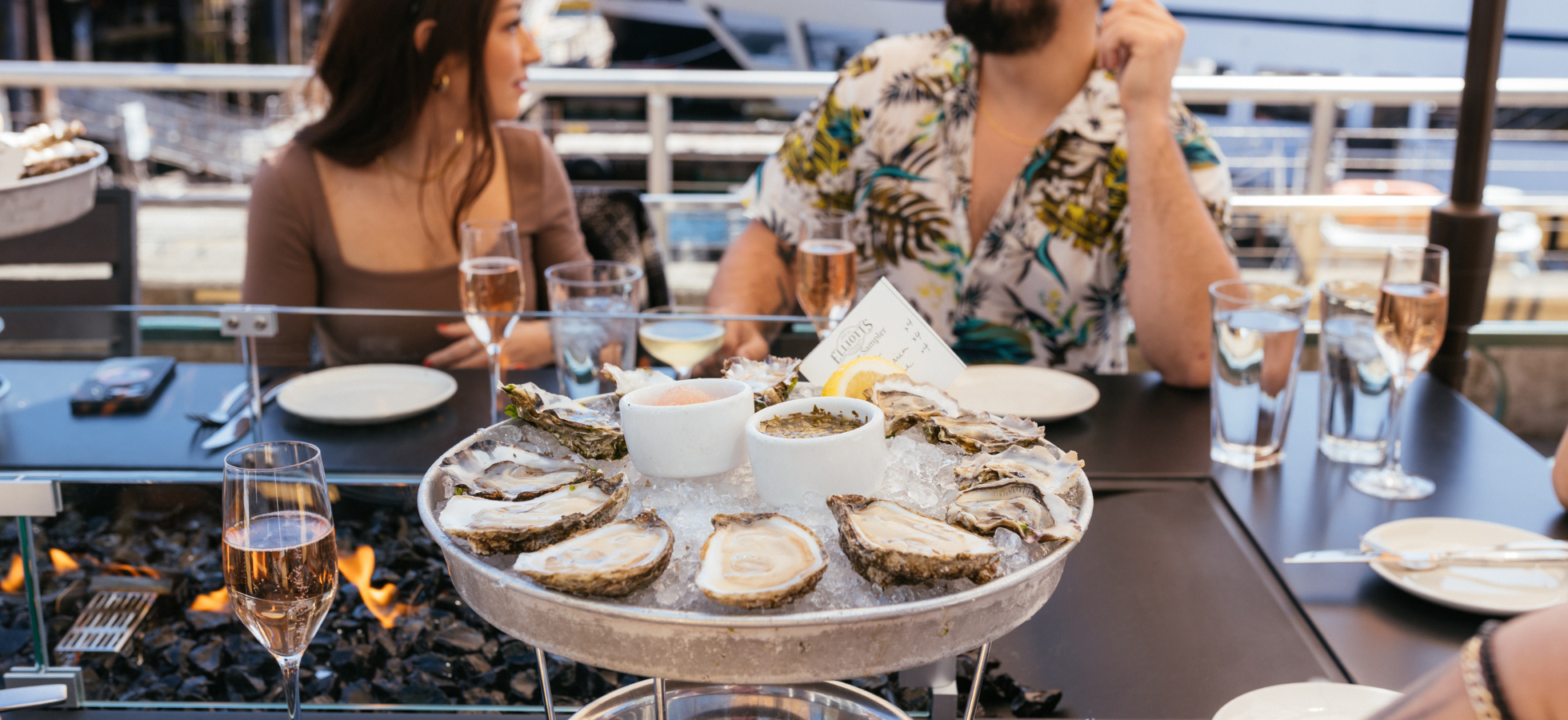 A large platter of oysters on ice is on a restaurant table, surrounded by glasses of wine and water. Two people sit across from each other, engaged in conversation at the waterfront.