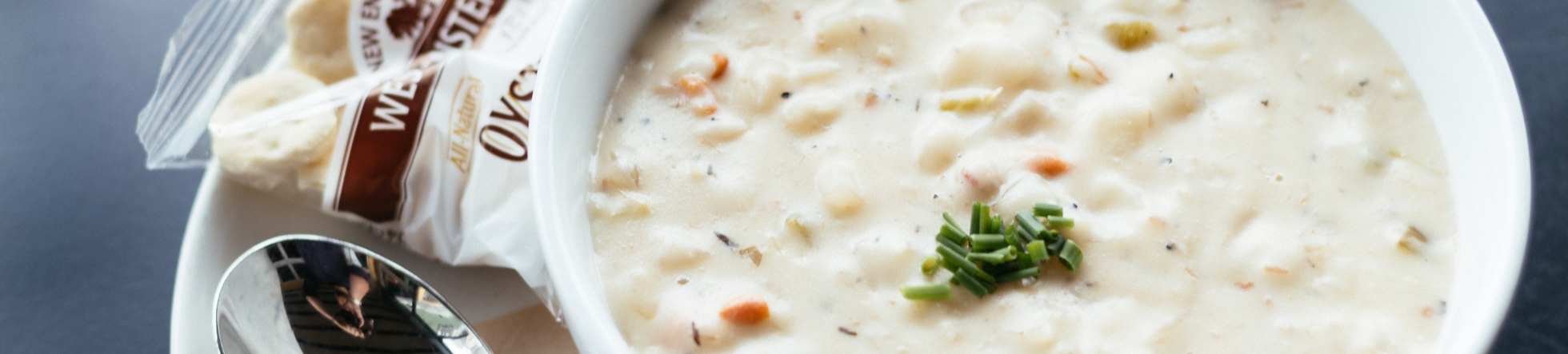 A bowl of creamy clam chowder garnished with chopped chives, served with a spoon and a packet of oyster crackers on a white plate.