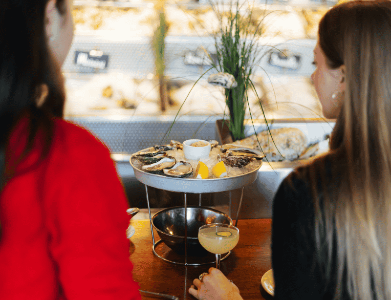 Two women sit at a table in a restaurant, facing a large seafood platter with oysters, lemon wedges, and dipping sauce. A cocktail and bowl are on the wooden table in front of them.