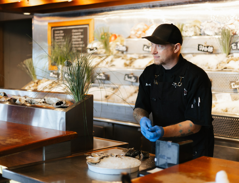 A seafood chef wearing a black uniform and blue gloves stands behind a counter shucking oysters, with fresh seafood displayed on ice in the background.