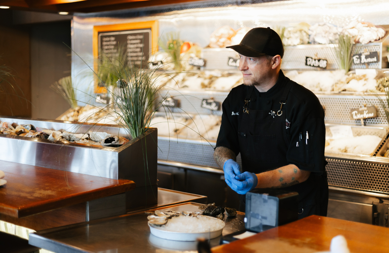 A seafood chef wearing a black uniform and blue gloves stands behind a counter, preparing oysters in a seafood market with various shellfish displayed on ice in the background.