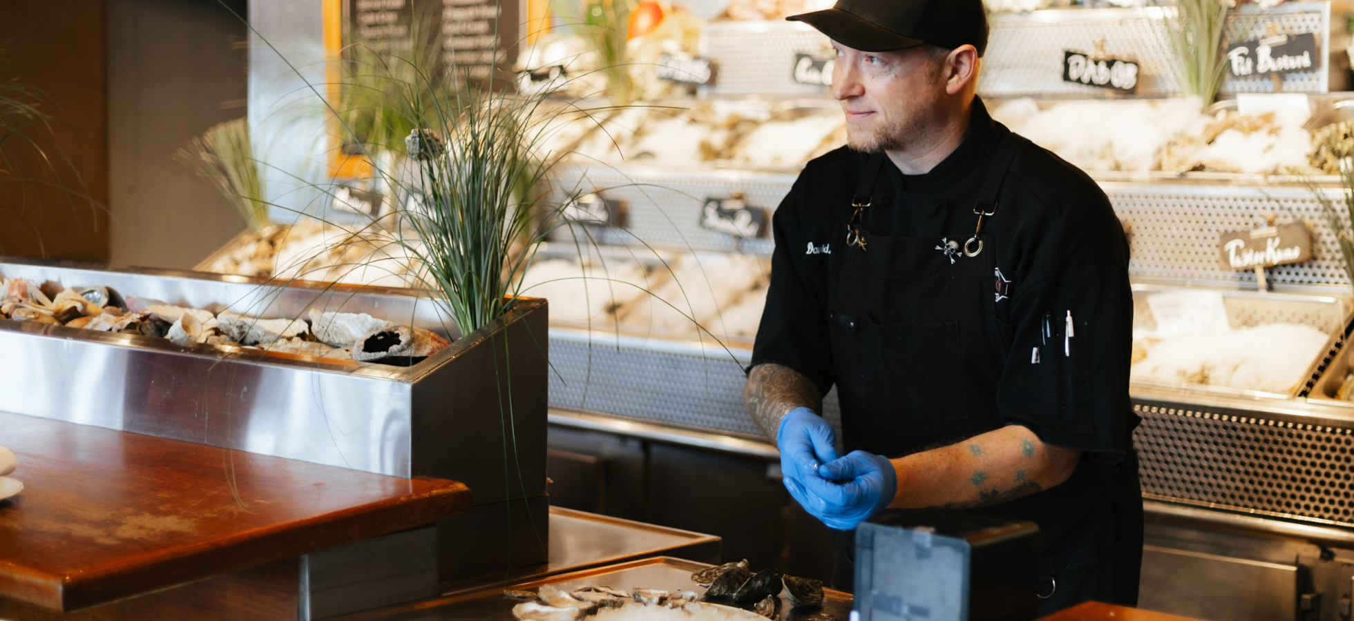 A person wearing a black chefs uniform and blue gloves stands behind a seafood counter displaying various fish and shellfish on ice.