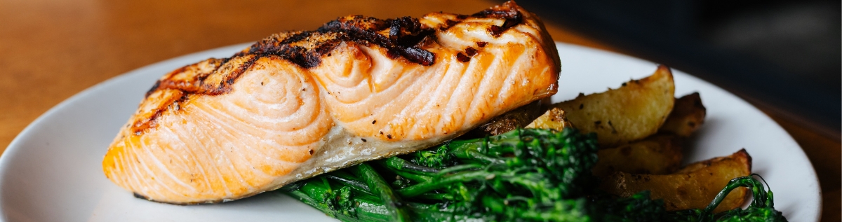 Grilled salmon fillet served on a white plate with roasted potato wedges and sautéed broccolini on a wooden table.