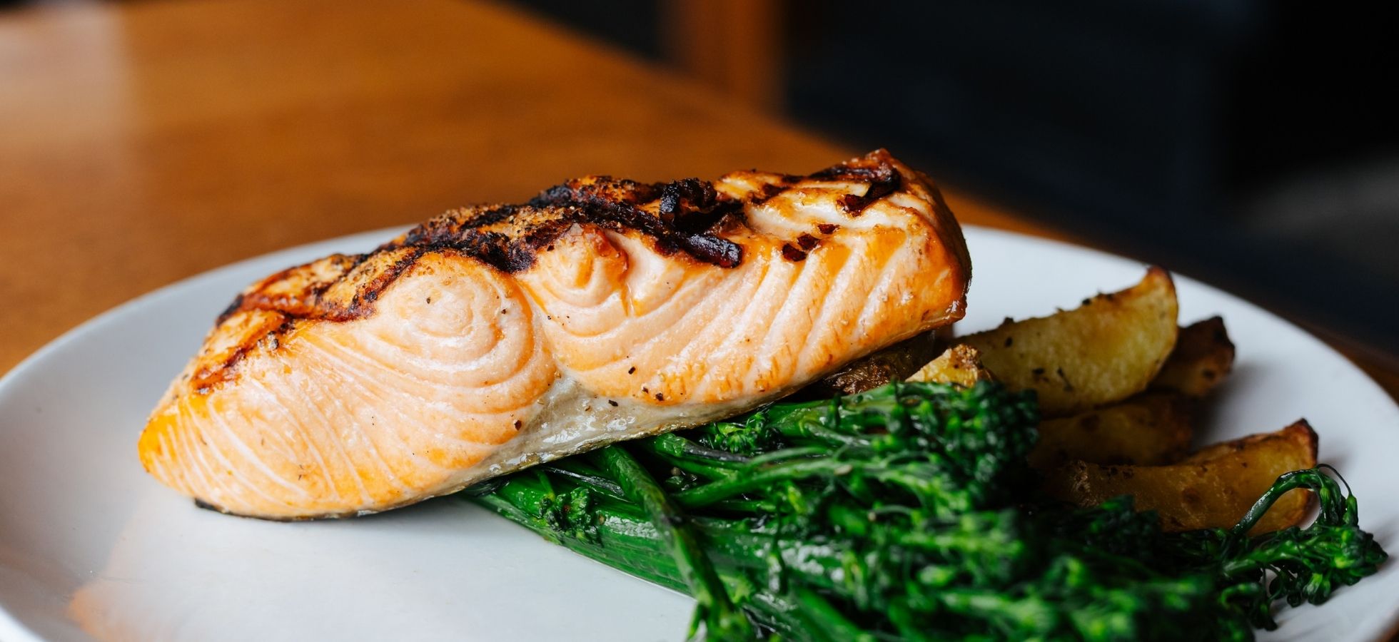 Grilled salmon fillet served on a white plate with roasted potato wedges and sautéed broccolini, placed on a wooden table.